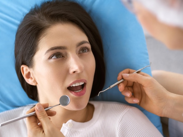 Mulher morena sorridente sendo examinada pelo dentista na clínica odontológica. Mãos de um médico segurando instrumentos odontológicos perto da boca do paciente. Dentes saudáveis e conceito de medicina.