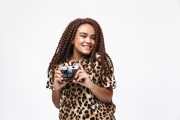 Mulher morena sorrindo e fotografando na câmera retro em pé, isolado contra uma parede branca