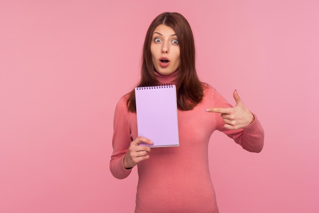 Mulher morena surpreendida em suéter rosa apontando o dedo para o caderno vazio em branco olhando para a câmera com expressão chocada espaço publicitário tiro de estúdio interior isolado no fundo rosa