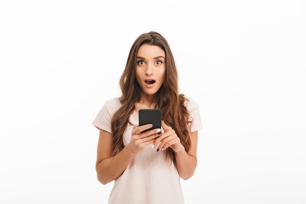 Foto mulher morena surpresa no t-shirt furando o smartphone com a boca aberta sobre parede branca