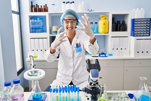 Mulher morena trabalhando no laboratório de cientistas gritando frustrada de raiva, mãos tentando estrangular, gritando loucamente