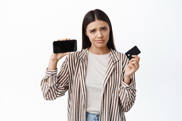 Mulher morena triste e desapontada mostrando a tela do smartphone e um cartão de crédito de plástico, carrancuda e com uma careta chateada, em pé sobre uma parede branca