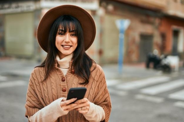 Mulher morena usando chapéu de inverno sorrindo usando smartphone ao ar livre na cidade