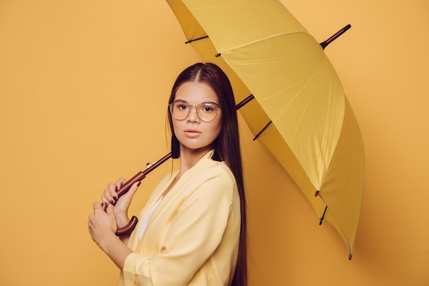 Mulher moreno nova nos vidros que vestem o revestimento amarelo que guarda o guarda-chuva amarelo sobre o fundo amarelo do estúdio.