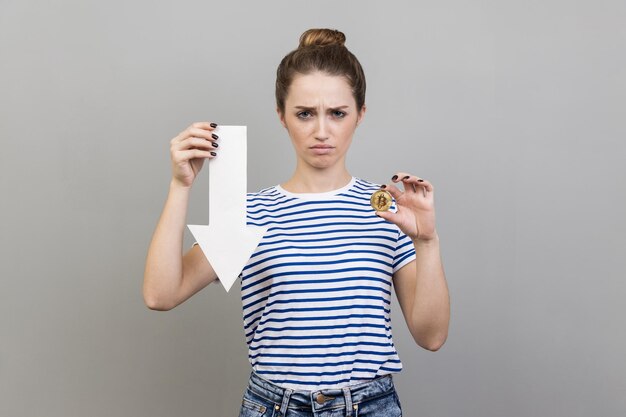 Foto mulher mostrando bitcoin e flecha branca apontando para baixo expressando emoções negativas