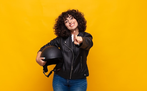 mulher motociclista sorrindo com orgulho e confiança fazendo a pose número um triunfantemente, sentindo-se uma líder