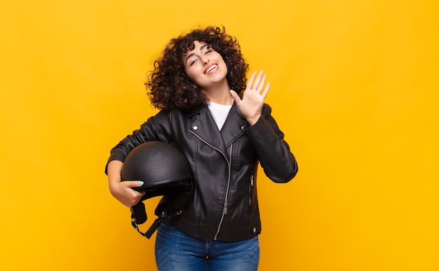 Mulher motociclista sorrindo feliz e alegremente, acenando com a mão, dando as boas-vindas e cumprimentando você ou dizendo adeus