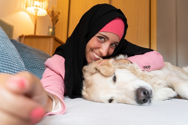 Mulher muçulmana alegre com animal de estimação usando um hijab dentro de casa. Vista lateral horizontal da mulher árabe deitada na cama com o cachorro em casa. Animais e estilo de vida das pessoas.