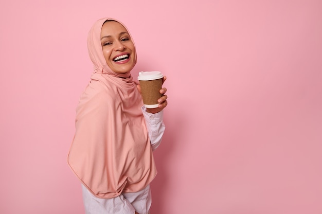 Mulher muçulmana árabe com a cabeça coberta por um hijab rosa segura um copo descartável de papelão para viagem, sorri um sorriso dentuço, olhando para a câmera, em pé contra um fundo colorido com espaço de cópia