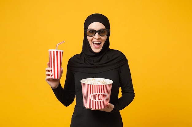 Mulher muçulmana árabe em roupas pretas de hijab óculos 3d imax assistir filme filme segurar pipoca, copo de refrigerante isolado no retrato de parede amarela. Conceito de estilo de vida de pessoas. .