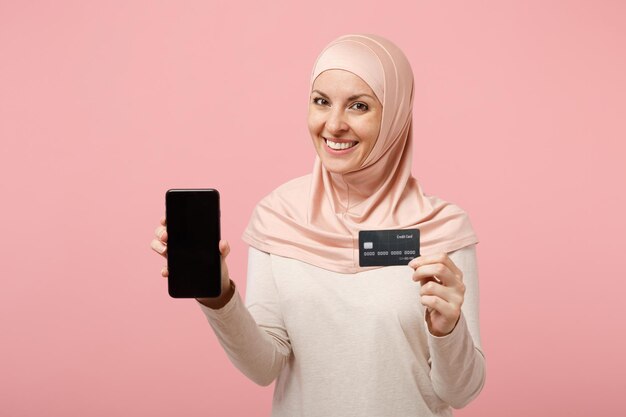 Mulher muçulmana árabe sorridente em roupas leves hijab posando isolado no fundo rosa. Conceito de estilo de vida religioso de pessoas. Mock up espaço de cópia. Segure o celular com o cartão de crédito de tela vazia em branco.