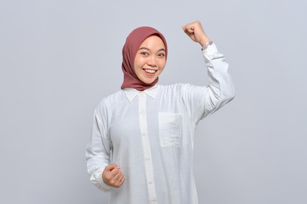 Mulher muçulmana asiática animada levantando os punhos comemorando a vitória com expressão facial eufórica isolada sobre fundo branco