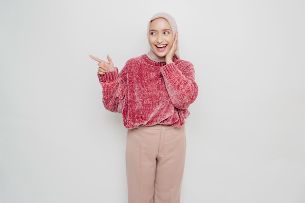 Mulher muçulmana asiática animada vestindo suéter rosa e hijab apontando para o espaço da cópia ao lado dela isolado por fundo branco