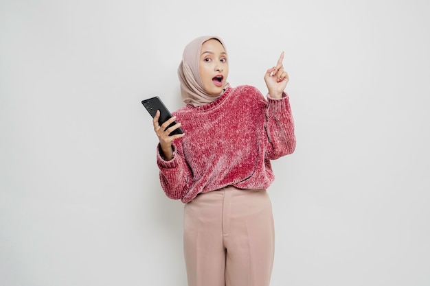 Mulher muçulmana asiática chocada vestindo suéter rosa e hijab apontando para o espaço da cópia em cima dela enquanto segura o telefone isolado pelo fundo branco