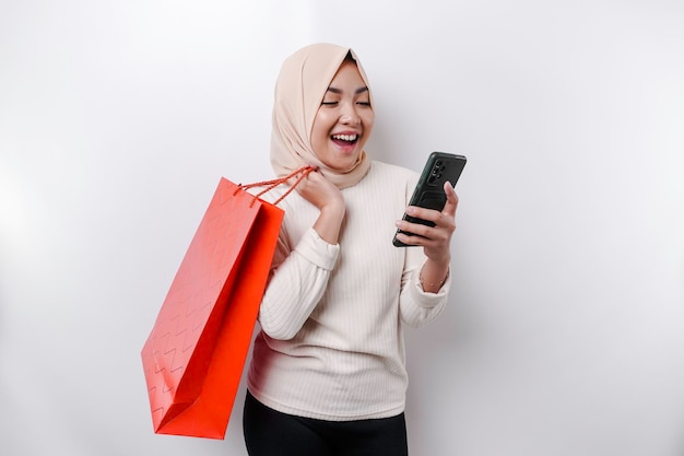 Mulher muçulmana asiática de retrato animada segurando uma sacola de compras on-line e sua foto de estúdio de smartphone isolada em fundo branco