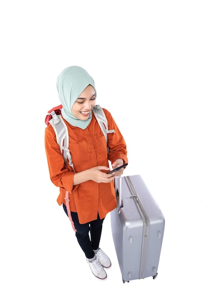 Mulher muçulmana asiática em um lenço na cabeça com uma mala e mochila segurando um telefone celular