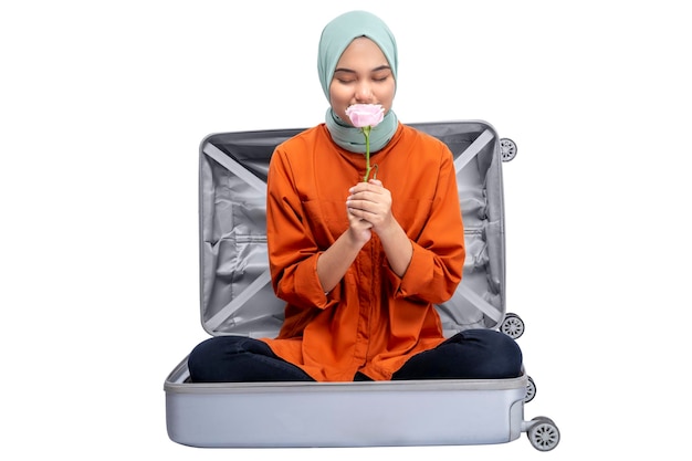Mulher muçulmana asiática em um lenço na cabeça sentado em uma mala enquanto segura uma flor rosa