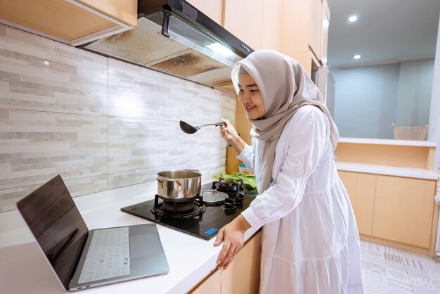 Mulher muçulmana asiática feliz olhando para seu laptop aprendendo a cozinhar um prato em casa