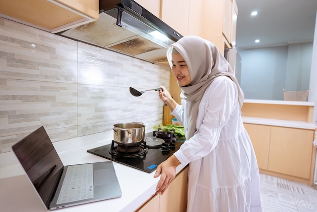 Mulher muçulmana asiática feliz olhando para seu laptop aprendendo a cozinhar um prato em casa