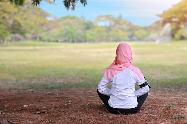 Mulher muçulmana asiática nova da vista traseira que senta-se na grama, apreciando a meditação. Garota pacífica no sportswear com hijab rosa pratica ioga em árvores de natureza verde com paz e tranquilidade. Conceito saudável e esporte