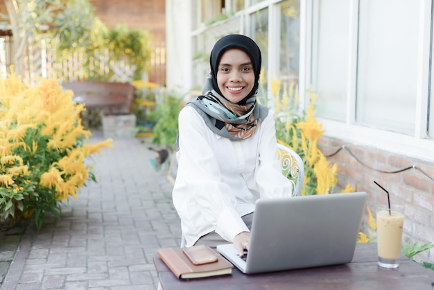 Mulher muçulmana asiática senta-se em um jardim com um laptop e um telefone no colo