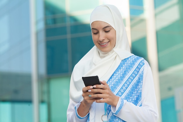 Mulher muçulmana bonita que texting em um telefone móvel ao ar livre.