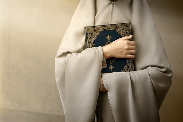 Mulher muçulmana com um véu de pé e segurando o Alcorão com fundo de parede cinza