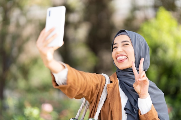 Mulher muçulmana contando uma selfie enquanto gesticula vitória com os dedos