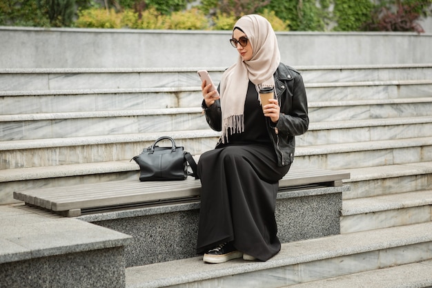 Mulher muçulmana elegante e moderna em hijab, jaqueta de couro e abaya preta sentada na rua da cidade com o celular em óculos de sol