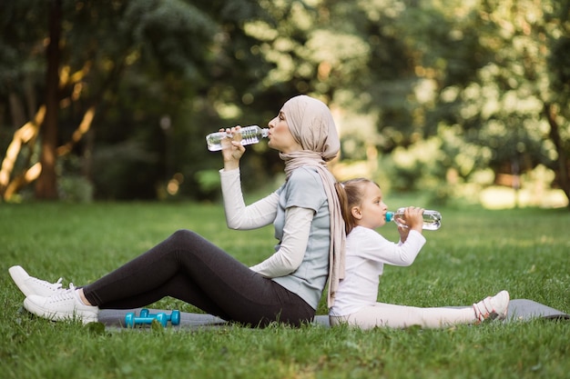 Mulher muçulmana em hijab sentada na esteira com sua filha no parque e água potável