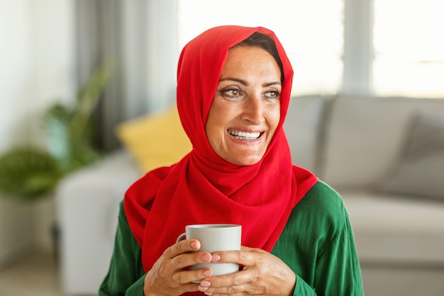 Mulher muçulmana madura feliz sonhando bebendo café e pensando em algo olhando de lado sentado na sala de estar