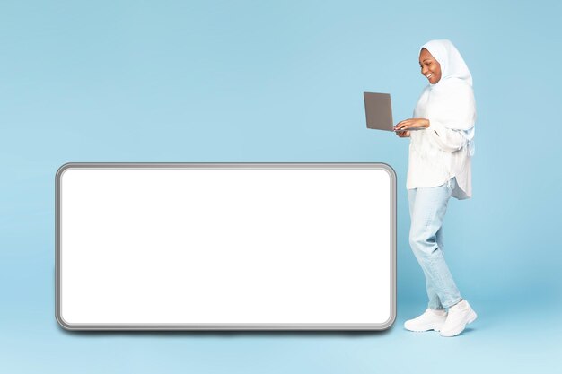 Foto mulher muçulmana negra feliz com laptop parado perto de um celular enorme com maquete de tela vazia branca