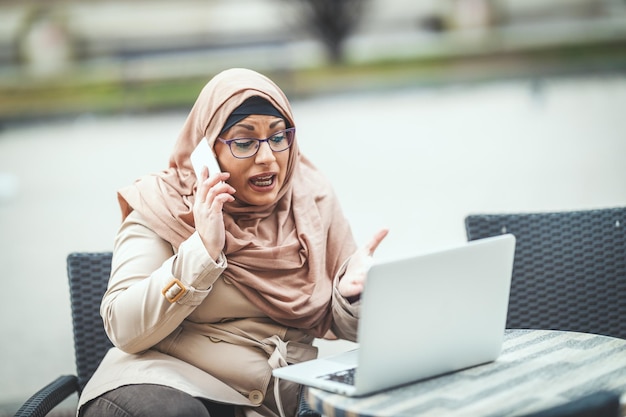 Mulher muçulmana preocupada de meia idade usando hijab está sentada no café em ambiente urbano, falando pelo smartphone e trabalhando no laptop.