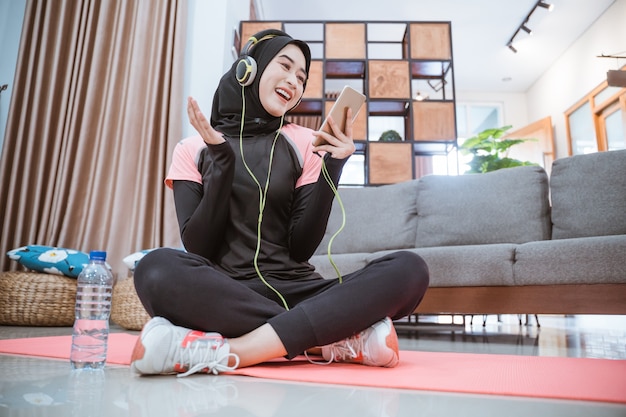 Mulher muçulmana vestindo uma roupa esportiva hijab usando fones de ouvido e um celular enquanto ouve música está sentada em uma esteira de ioga