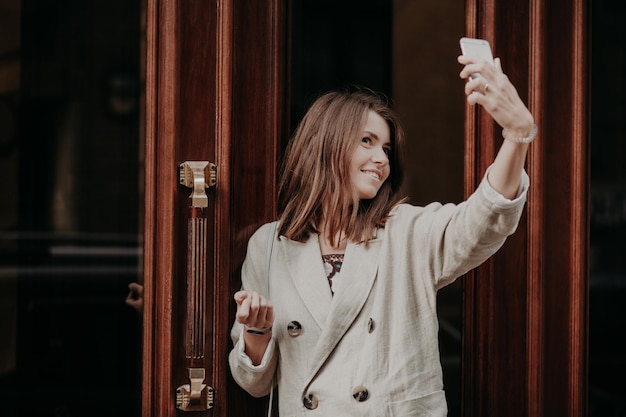 Mulher muito adorável faz selfie com telefone inteligente vestido com capa de chuva posa ao ar livre usa tecnologia moderna compartilha fotos em redes sociais Tecnologia de pessoas e conceito de entretenimento