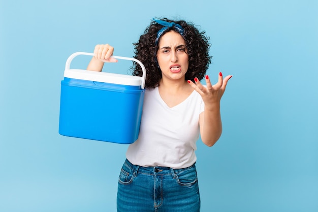 Mulher muito árabe olhando irritado irritado e frustrado e segurando uma geladeira portátil