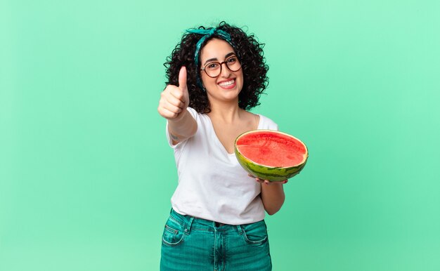 Foto mulher muito árabe, sentindo-se orgulhosa, sorrindo positivamente com os polegares para cima e segurando uma melancia. conceito de verão