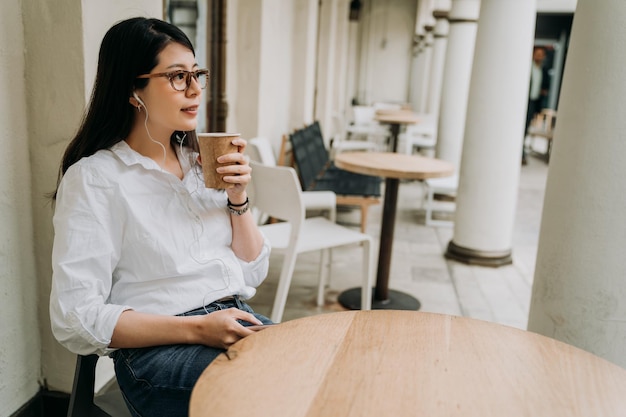 Mulher muito asiática no café da rua ao ar livre, sentado à mesa, ouve música em fones de ouvido. jovem segurando o celular relaxando no tempo livre. Escritório móvel no conceito de descanso de vida de verão.