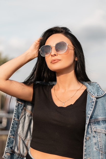 Mulher muito atraente jovem hippie no top preto com uma jaqueta jeans azul em elegantes óculos de sol pretos relaxa perto de uma parede de madeira vintage. Modelo de garota morena sexy com roupas da moda descansa ao ar livre.