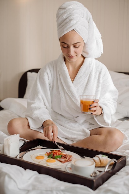 Mulher muito caucasiana em roupão de banho e toalha na cabeça tomando café da manhã na cama de manhã no quarto de hotel