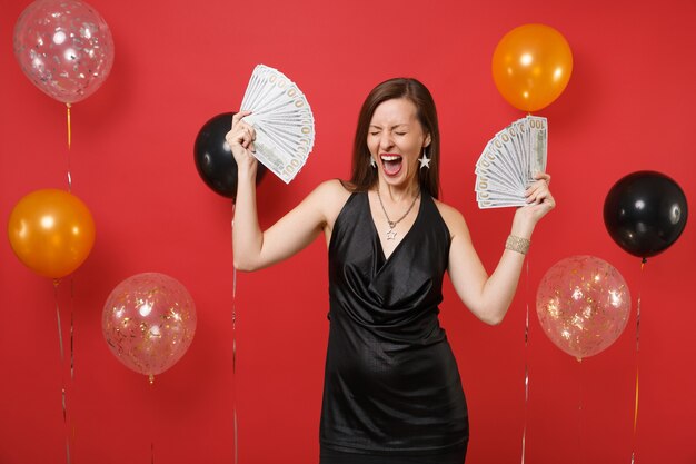 Mulher muito feliz em um vestido preto gritando, comemorando, segurando um pacote de muitos dólares, dinheiro em balões de ar de fundo vermelho brilhante. Feliz Ano Novo, conceito de festa de feriado de maquete de aniversário.