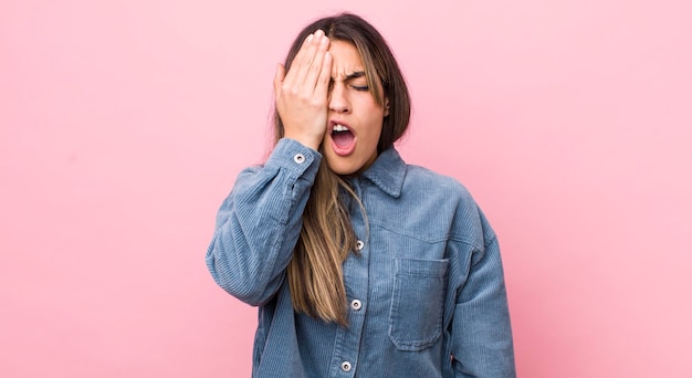 Mulher muito hispânica parecendo sonolenta entediada e bocejando com dor de cabeça e uma mão cobrindo metade do rosto