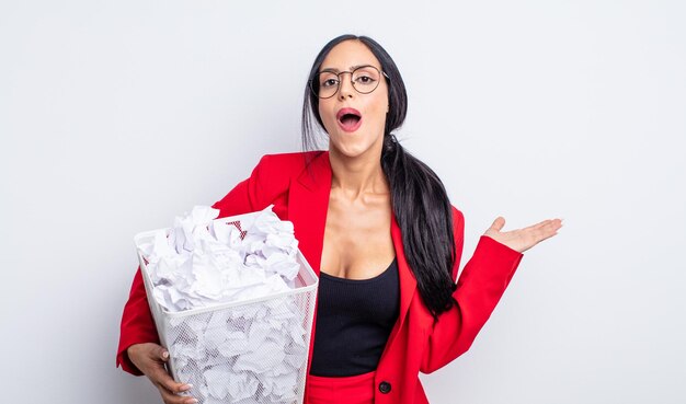 Mulher muito hispânica parecendo surpresa e chocada, com o queixo caído segurando um conceito de lixo de bolas de papel objeto