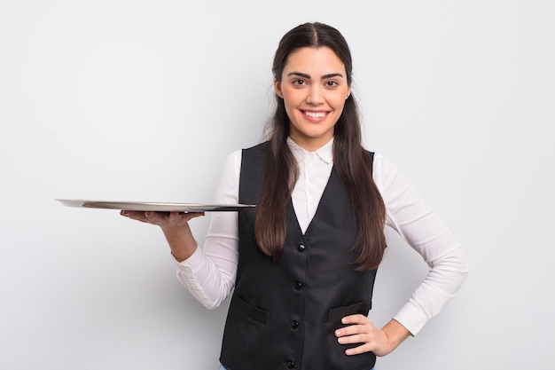 Mulher muito hispânica sorrindo alegremente com uma mão no quadril e garçom confiante com o conceito de bandeja