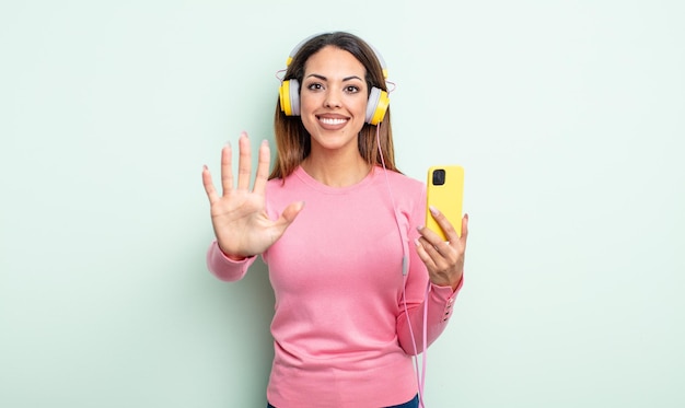 Mulher muito hispânica sorrindo e parecendo amigável mostrando o conceito de smartphone e fones de ouvido número cinco