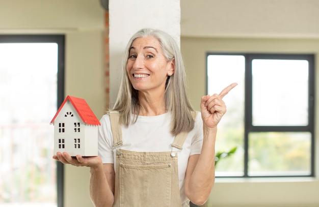 mulher muito idosa sorrindo alegremente, sentindo-se feliz e apontando para o lado com um modelo de casa