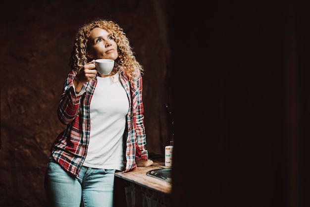 Mulher muito jovem de meia-idade em casa bebendo e olhando para cima pensando e curtindo relaxar sozinha uma mulher serena independente em atividade de lazer interna com uma xícara de café planejamento