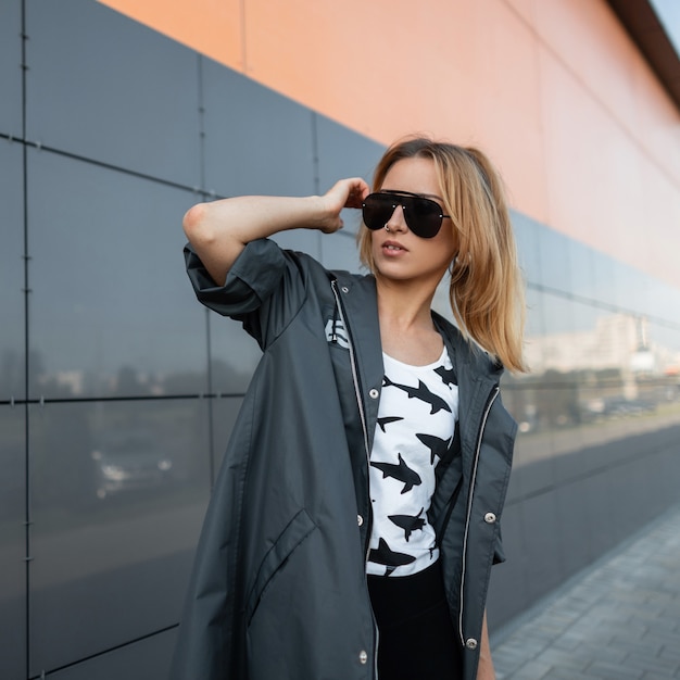 Mulher muito jovem e atraente com roupas elegantes em elegantes óculos de sol pretos posa em uma cidade perto de um edifício moderno e cinza. garota ruiva europeia gosta de um dia ensolarado.