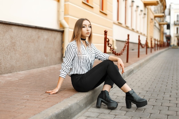 Mulher muito jovem e bonita em uma elegante blusa listrada preto e branca em jeans da moda em sapatos de couro com um piercing no nariz, descansando sentado ao ar livre na cidade.