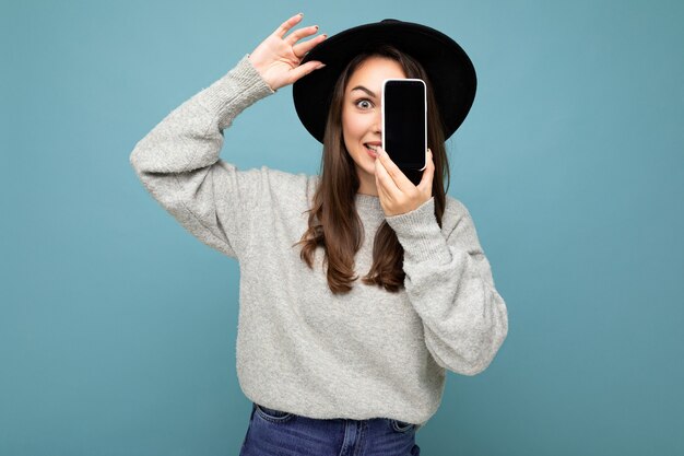 Mulher muito jovem e sorridente com chapéu preto e suéter cinza segurando o telefone